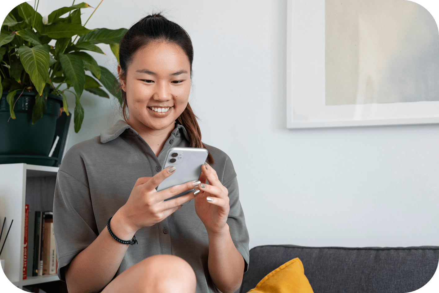 Smiling woman sitting on couch using smartphone in modern living room with potted plant, bookshelf, and abstract wall art