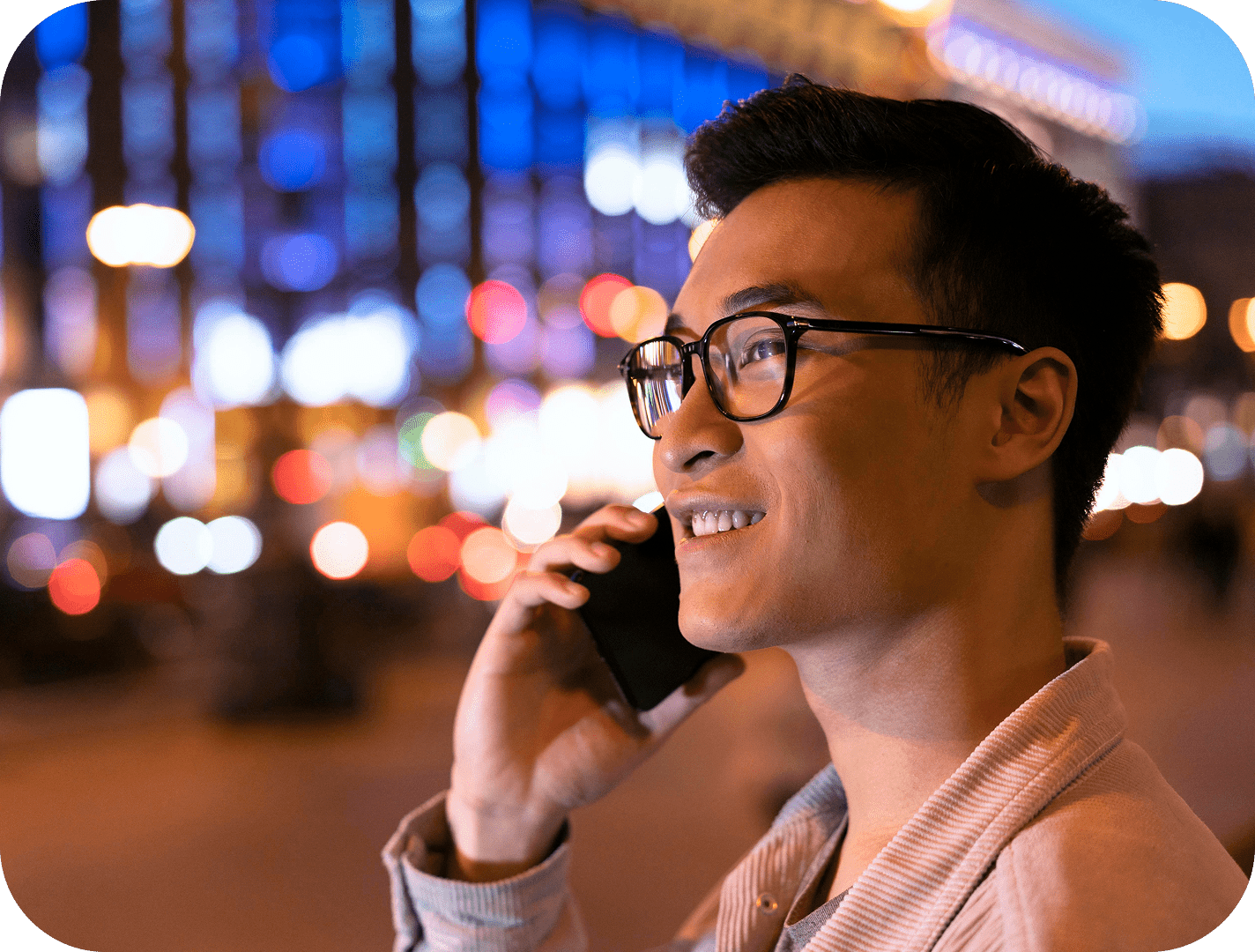Smiling man wearing glasses talking on smartphone at night with colorful city lights blurred in background