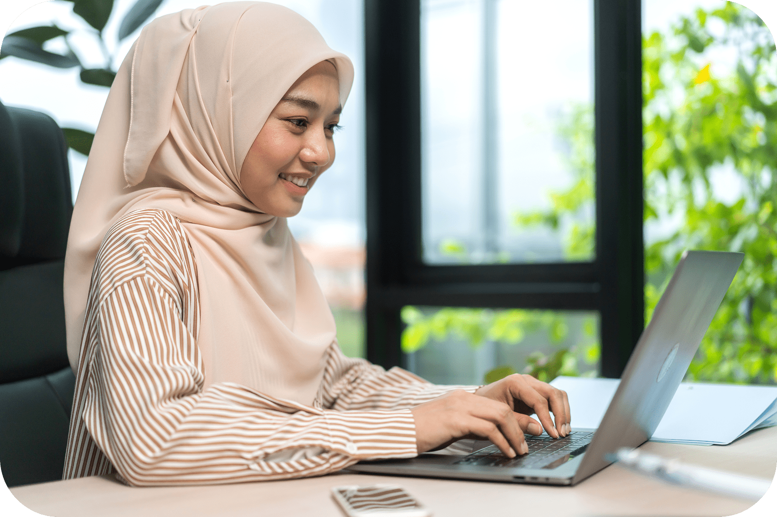 A smiling woman in a beige hijab and striped shirt works on a laptop at a desk. Sunlight filters through a window with greenery outside.