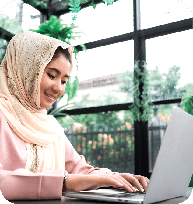 Smiling woman in a hijab types on a laptop in a bright, plant-filled café with large windows, conveying a sense of focus and positivity