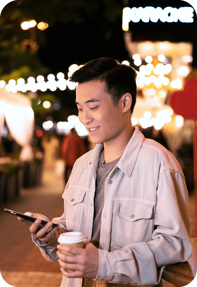 Smiling person with coffee checking phone at night, festive lights behind