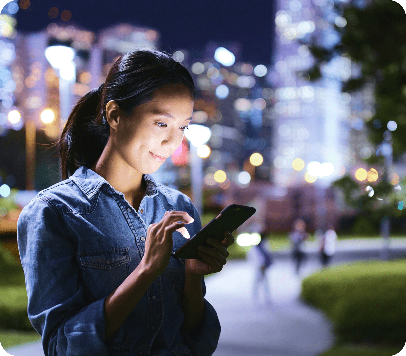Woman in denim shirt smiling at her phonein night street with blurred city lights behind